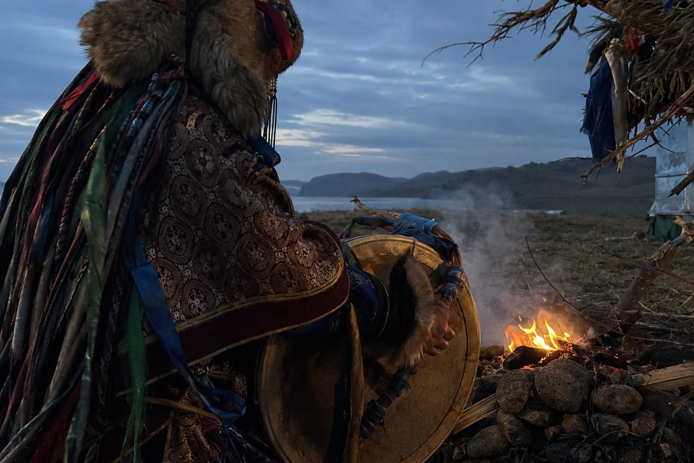 A shaman seated on the shore of Lake Baikal at dusk, holding his drum over the flames, smoke rising past a ribboned post