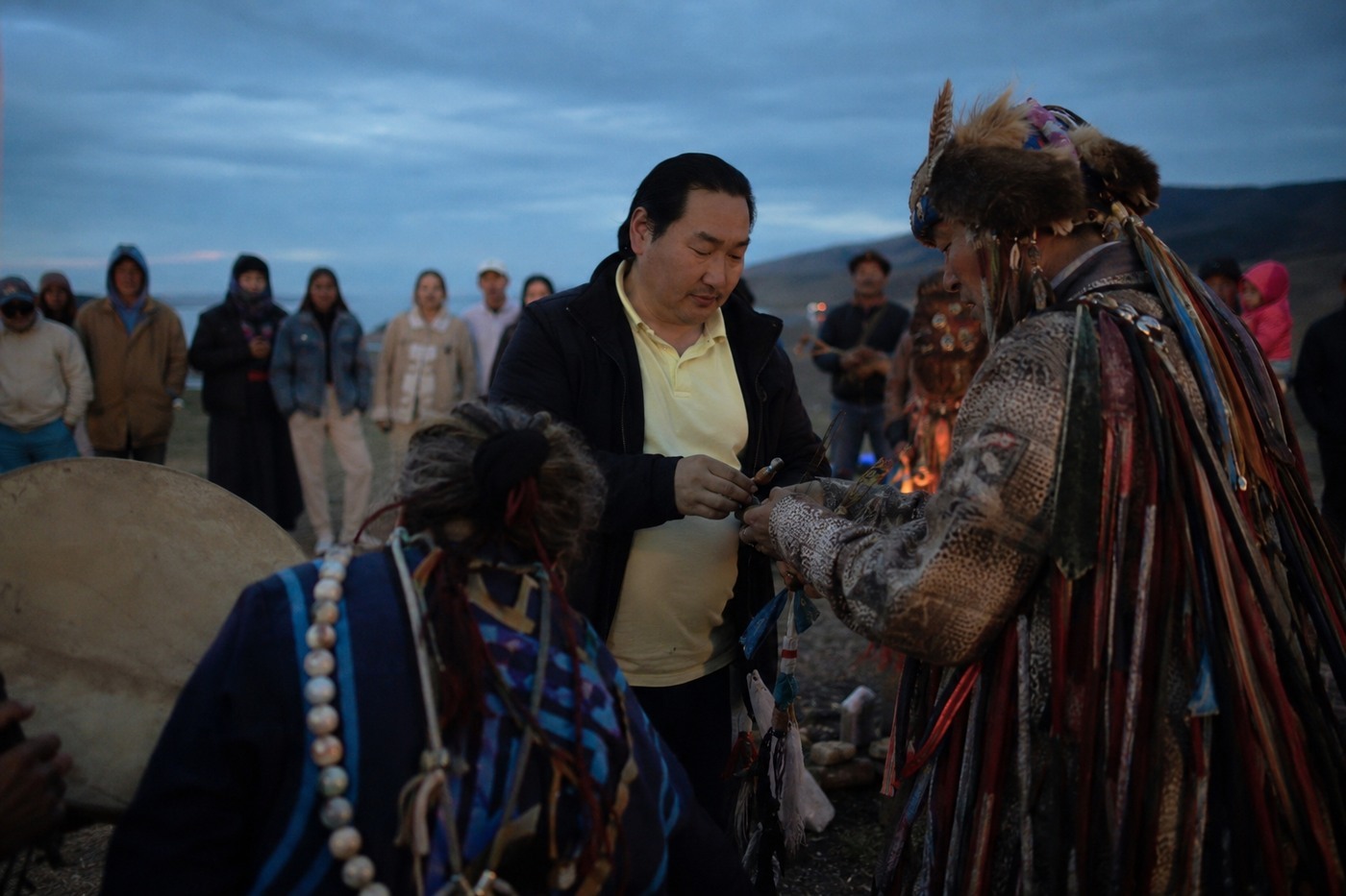 A shaman in ceremonial dress placing something into the cupped hands of a man standing before him, the circle watching and a fire burning behind