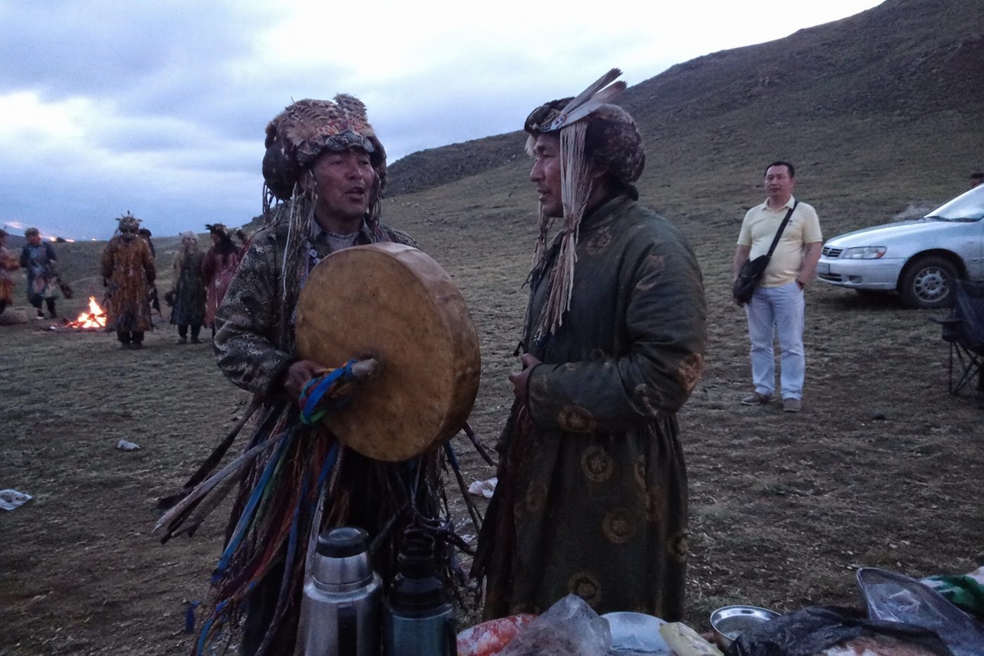 Two shamans in ribboned coats and feathered headdresses talking beside a drum at dusk, the ceremonial fire burning behind them