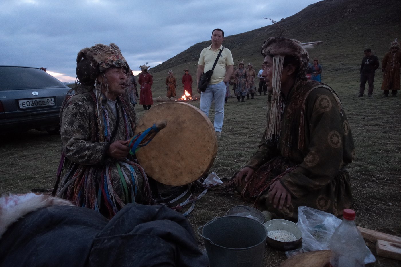 Two shamans sitting together on the grass after the ceremony, one holding a drum, speaking while others stand near the fire