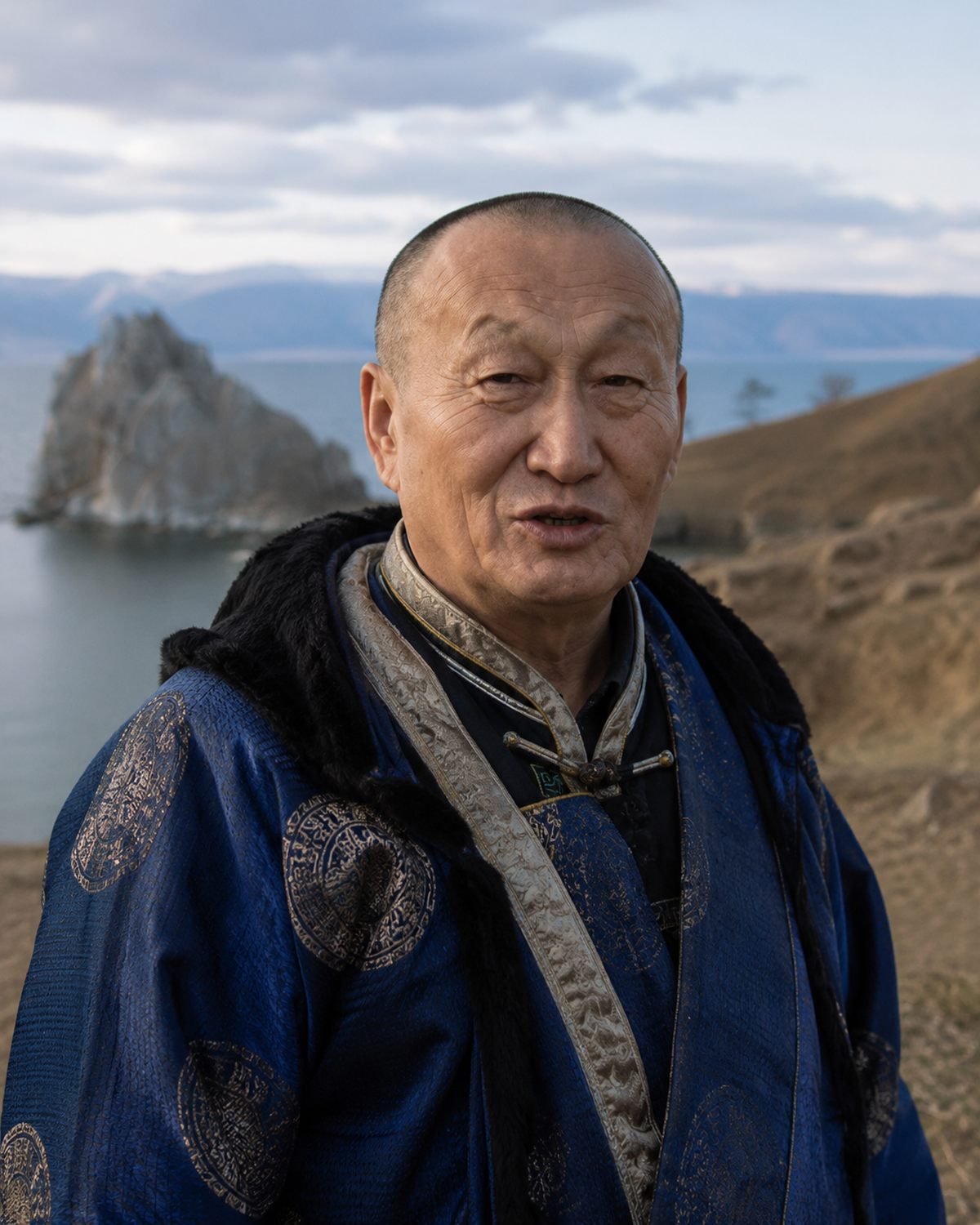 Bair Zhambalovich Tsyrendorzhiev in a blue silk deel on the headland at Cape Burkhan, Shaman Rock rising from Lake Baikal behind him